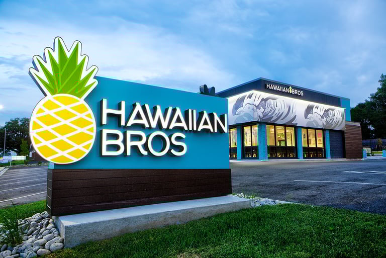 Hawaiian Bros storefront with large pineapple logo sign and modern teal and brown building exterior at dusk