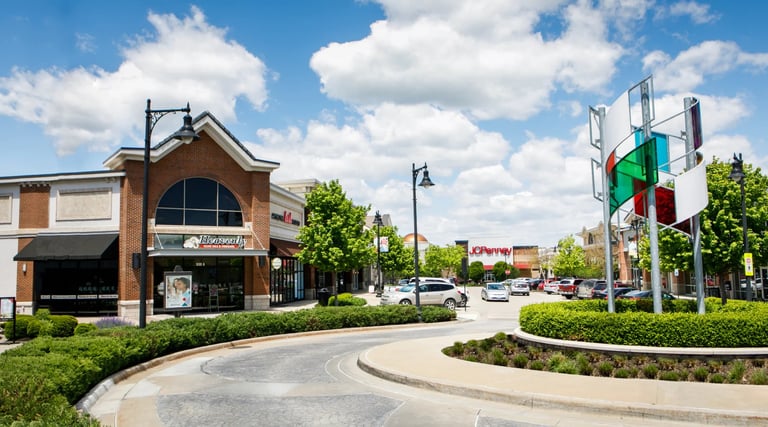 Suburban shopping center with brick storefront building, curved driveway, landscaping, and directional signage under blue sky