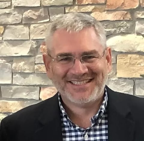 Smiling man with gray hair and glasses wearing a black blazer and checkered shirt in front of a stone wall